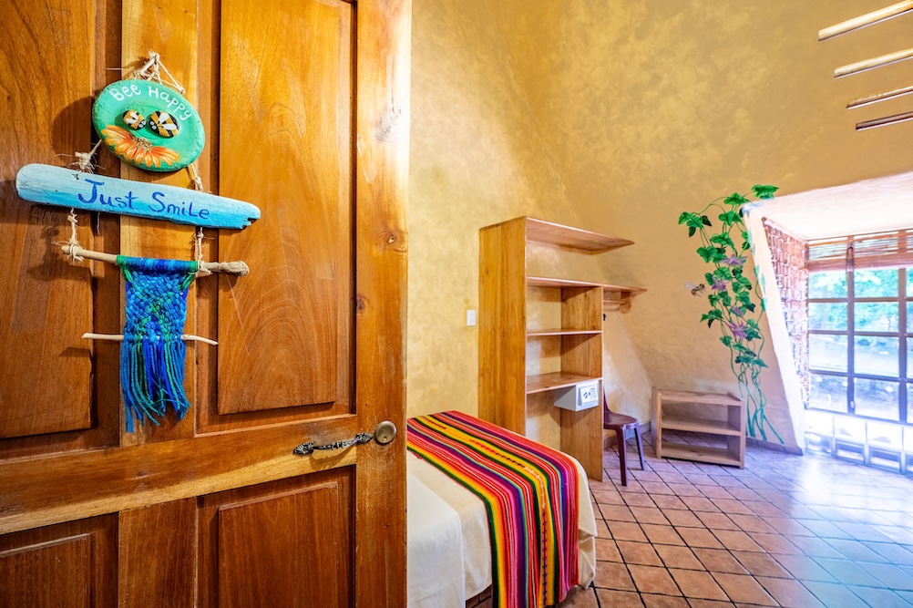 A cozy room with yellow walls, a single bed covered with a colorful striped blanket, wooden shelves, a chair, and window. The open door has a decorative sign that says “Just Smile.”.