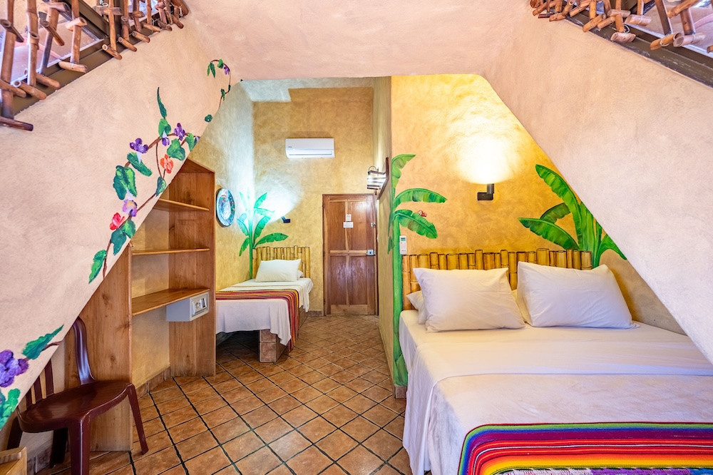 A hotel room with two beds, colorful blankets, decorative wall paintings of leaves and flowers, tiled floor, wooden furniture, and a wall-mounted air conditioner above the door.