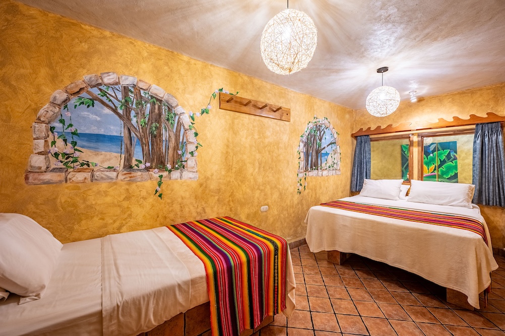 A cozy hotel room with a double bed and a twin bed, each with white bedding and colorful striped blankets. The walls are painted yellow with beach mural art and arched window frames. The floor is tiled.