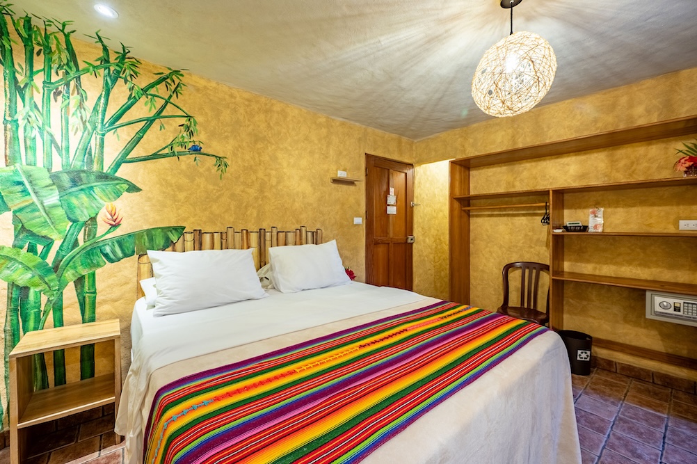 A cozy hotel room with a double bed, white linens, and a colorful striped blanket. The walls are painted yellow, one featuring a mural of green bamboo. There are shelves, a chair, and a woven light fixture.