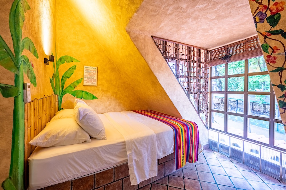 A small bedroom features a single bed with white linens and a colorful striped blanket. The walls are painted yellow with a green plant mural, and large windows with bamboo blinds let in natural light.